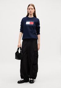 Felpa navy con logo bianco e rosso, pantaloni cargo neri con tasche, borsa nera con manici corti e scarpe nere con suola spessa.