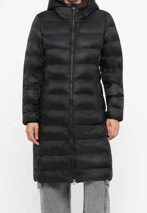 Femme portant un long manteau noir matelassé à capuche avec fermeture éclair devant et poches latérales, associé à un jean gris clair.