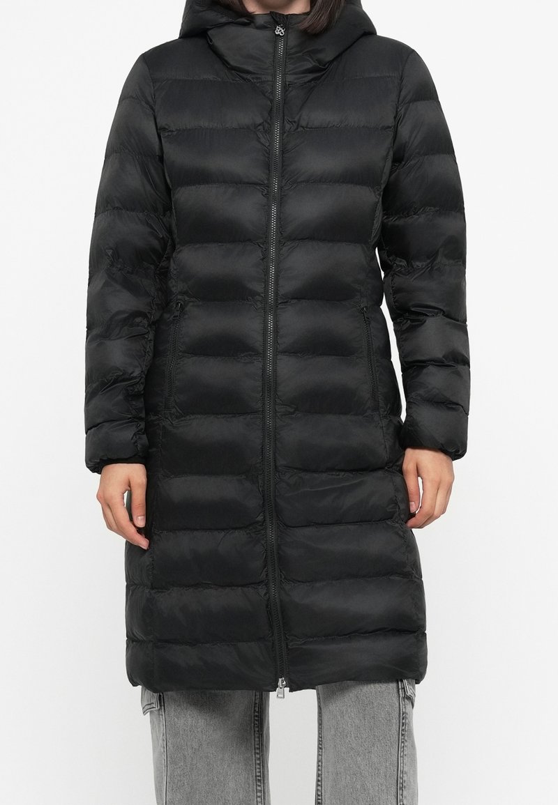 Femme portant un long manteau noir matelassé à capuche avec fermeture éclair devant et poches latérales, associé à un jean gris clair.