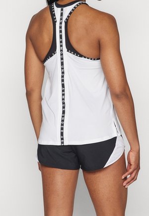 Kvinde iført hvid racerback tanktop med sort Under Armour-logobånd og sorte sportsshorts, set bagfra.