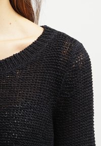 Svart strikket genser med ribbestrikket hals, med en åpen vevstruktur og en avslappet passform. Stoffet virker mykt og pustende.