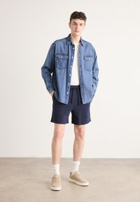 Camisa de denim con dos bolsillos en el pecho, color azul claro, combinada con pantalones cortos azul marino. El modelo lleva zapatillas beige y calcetines blancos.