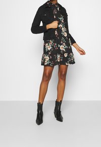 Schwarze Jeansjacke über einem schwarzen Blumenkleid mit gerüschtem Saum. Das Kleid hat mehrfarbige Blumen und wird mit schwarzen Stiefeletten kombiniert.