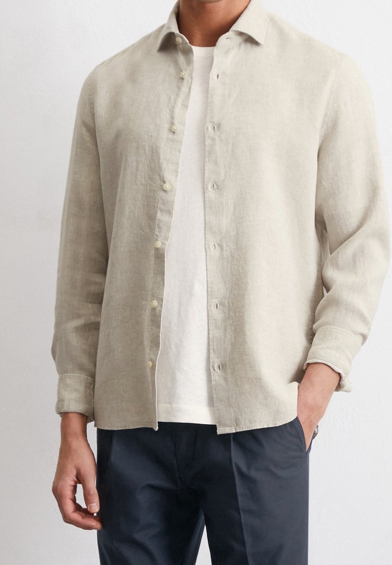 Homme portant une chemise en lin beige boutonnée sur un t-shirt blanc et un pantalon foncé, une main dans la poche, sur fond uni.