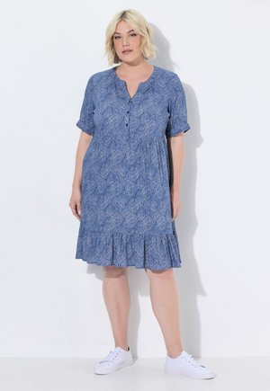 Kobieta w niebieskiej, krótkorękawowej sukience do kolan z subtelnym liściastym wzorem i białych trampkach, stojąca na tle jednolitej białej ściany.