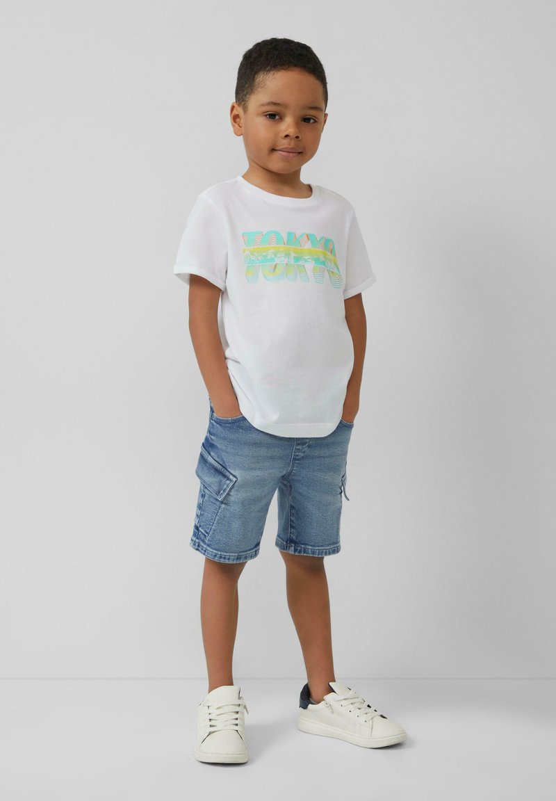 Witte katoenen T-shirt met kleurraf "TOKYO" grafiek, gecombineerd met denimshorts met zijzakken, en witte sneakers met zwarte accenten.