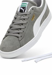 Pilki suede sportbačiai su baltais akcentais ir guminiais padais. Ypatybės: auksinis Puma logotipas šone, plokščios pilkos raišteliai ir paminkštinta liežuvėlis.