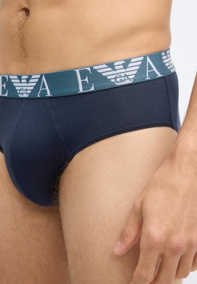 Cuecas de algodão azul-marinho com uma cintura elástica que apresenta um logótipo em tons de verde-azulado e branco, com textura suave e ajuste justo, mostrando parcialmente a coxa e a mão.