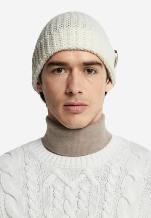 Hvid strikket hue med en struktureret overflade og en sølvdetalje. Båret med en lys brun turtle neck og en hvid kabelstrikket sweater.