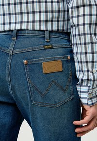 Man in blue plaid shirt wearing blue Wrangler jeans with leather patch and logo on back pocket, hand resting near pocket.