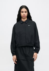 Μαύρο μπουφάν Nike με γιακά και cropped σχεδιασμό. Διαθέτει ελαστικά μανίκια και κάτω μέρος, πλαϊνές τσέπες και μικρό λευκό λογότυπο στο στήθος.
