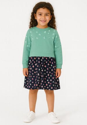 Junges Mädchen mit lockigem Haar, das einen grünen Sweatshirt mit Punkten und einen Navyrock mit bunten Punkten trägt, lächelt und steht vor einem weißen Hintergrund.