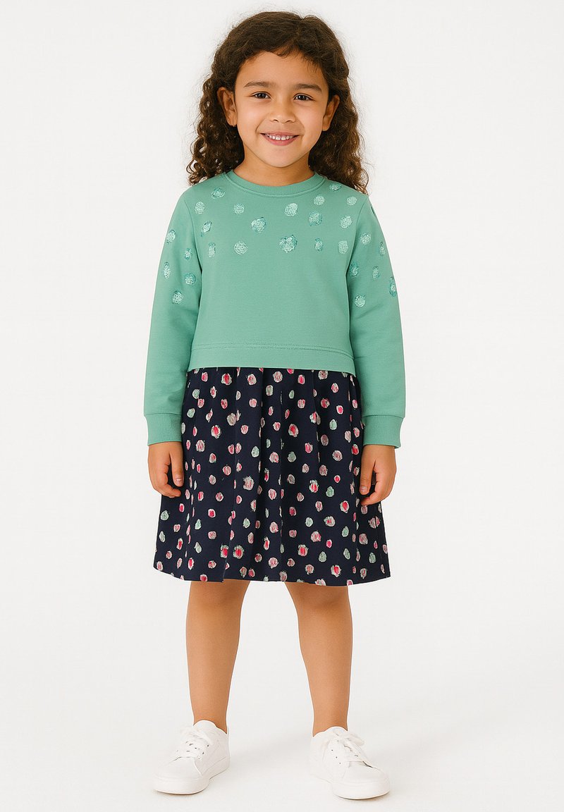 Junges Mädchen mit lockigem Haar, das einen grünen Sweatshirt mit Punkten und einen Navyrock mit bunten Punkten trägt, lächelt und steht vor einem weißen Hintergrund.