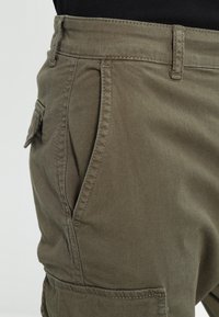 Gros plan d'une personne portant un pantalon cargo vert olive avec des poches latérales et à rabat ainsi que des détails de couture visibles.