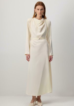 Vörös hajú nő hosszú, krém színű selyemruhába öltözve, amelynek drapált nyaka, öve, hosszú ujjai és bézs színű magassarkú cipője van, egy sima fal előtt áll.