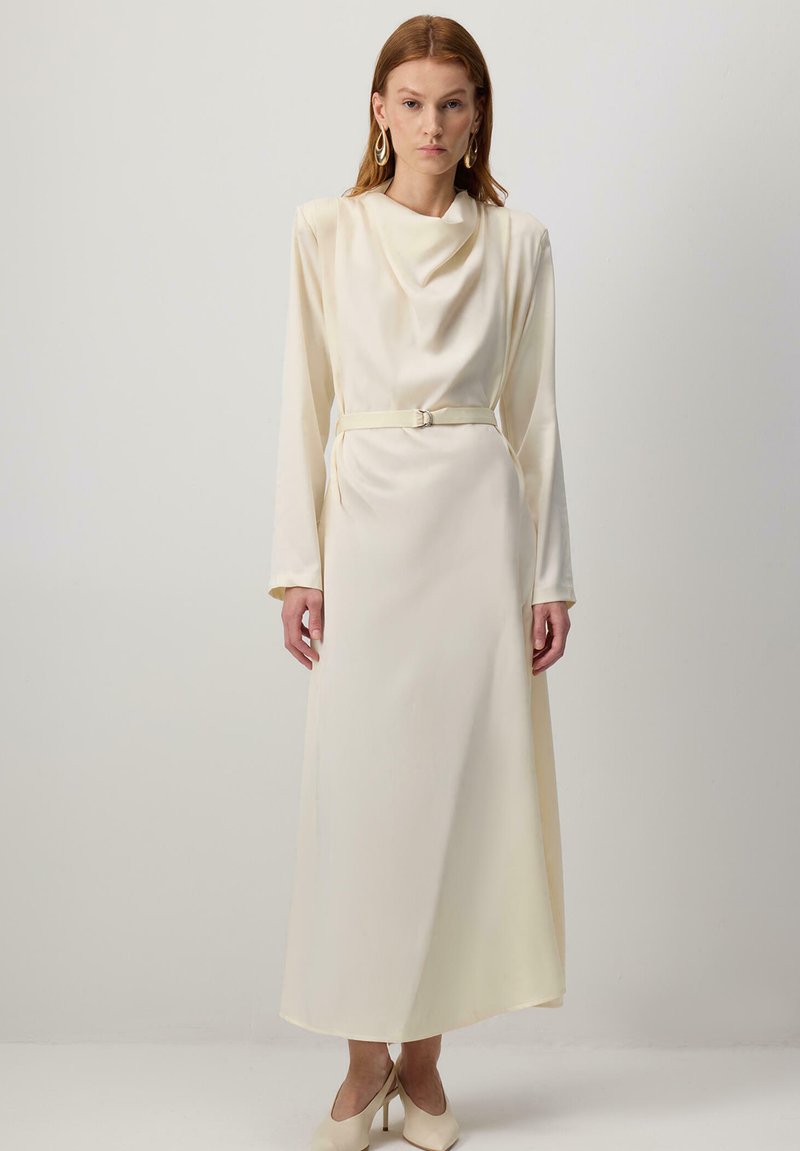 Punane juustega naine, kes kannab pikka kreemjat satiinist kleiti drapeeritud dekolteega, vöökoht, pikkade varrukatega ja beeže kõrge kontsaga kingi, seisab ühtlase seina ees.