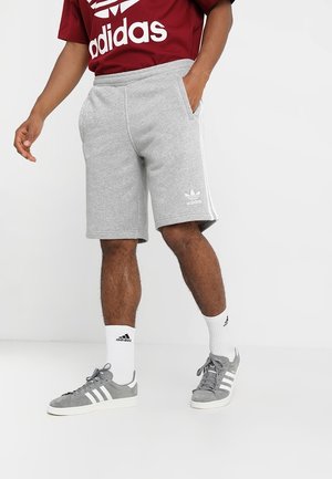 Mężczyzna ubrany w szare szorty Adidas, szare buty sportowe Adidas z białymi paskami, białe skarpetki Adidas oraz czerwoną koszulkę Adidas.