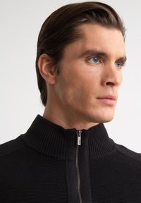 Sudadera negra con cremallera y cuello acanalado. Presenta un cierre de cremallera plateado y una tela suave, diseñada para un ajuste ceñido.