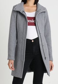 Cappotto di lana grigia con colletto alto, chiusura con zip e tasche laterali, abbinato a una camicia bianca e jeans neri. Texture liscia, vestibilità su misura.