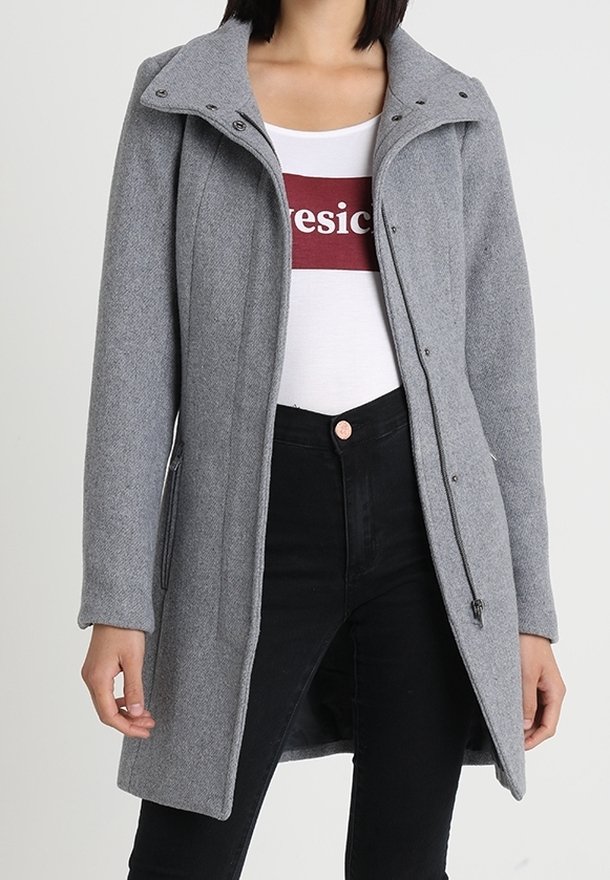 Cappotto di lana grigia con colletto alto, chiusura con zip e tasche laterali, abbinato a una camicia bianca e jeans neri. Texture liscia, vestibilità su misura.
