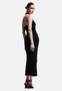 Musta halterneck-mekko, jossa on tiukka siluetti, keskivartalon pituus ja teksturoitu kangas. Yhdistetty mustiin hihnakenkiin.