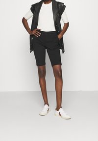 Černé na míru šité šortky, bílá žebrovaná košile a černá kožená vesta bez rukávů. Bílé tenisky s vínovými akcenty a jemnými detaily.