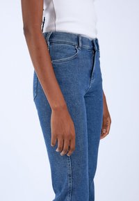 Jeans in denim blu con vita alta, vestibilità slim e dettagli di cucitura visibili. Il materiale sembra resistente con una texture liscia.