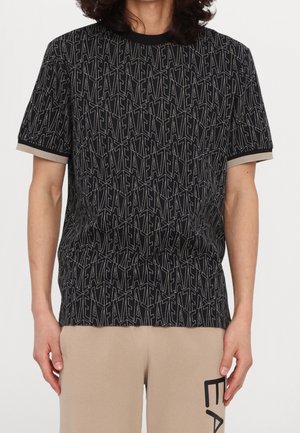 Homme portant un T-shirt noir à motifs avec le texte répété "EA7" et un short beige avec un logo partiel "EA7" visible.