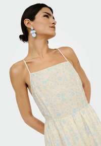 Robe à motifs floraux en beige clair avec des accents bleus. Fines bretelles spaghetti, buste ajusté et jupe fluide. Grandes boucles d'oreilles bleues remarquables.