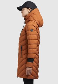 Vadderad lång kappa i orange med zigzag-quiltad design. Har huva, svarta muddar och fickor med dragkedja. Svart keps bärs.
