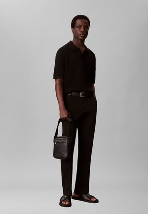 Hombre de pie frente a un fondo gris, lleva una camiseta negra de manga corta, pantalones negros, sandalias negras y sostiene un bolso negro de bandolera.