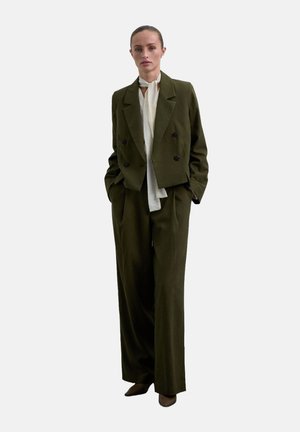 Chaqueta doble faz de color verde oliva con botones negros, combinada con pantalones de pierna ancha y una blusa de color claro, de pie frente a un fondo blanco.