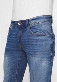 Jeans in denim blu scuro con una texture liscia, dotati di cinque tasche, chiusura con bottone in rame e dettagli di sfumatura discreta lungo le gambe.