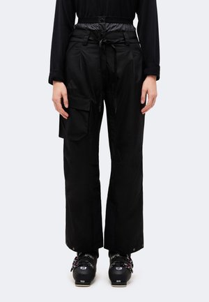Une personne porte un pantalon cargo noir à jambes larges avec une ceinture et des chaussures de ski noires, sur un fond uni.