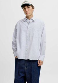 Ung mand iført en hvid skjorte med striber, mørkeblå jeans, hvid kasket og sølvfarvede ring-øreringe, som står foran en ensfarvet baggrund.