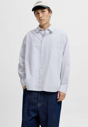 Junger Mann, der ein weißes gestreiftes Hemd mit Knopfleiste, dunkle Jeans, eine weiße Mütze und silberne Creolen trägt, steht vor einem schlichten Hintergrund.