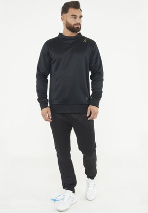 Man met baard, gekleed in een zwarte longsleeve en zwarte broek, loopt naar voren, witte sneakers, tegen een effen witte achtergrond.