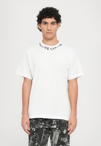 Balta trumpa rankovėmis marškinėliai su apvalia iškirpte, ant kurių yra juodas tekstas. Derinama su juodomis džinsais, puikuojančiomis detaliu baroko raštu.