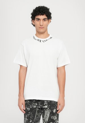 White short-sleeve T-shirt with a round neck featuring black text. Paired with black jeans displaying a detailed baroque pattern.