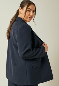 Granatowa marynarka wykonana z teksturowanej tkaniny, z klapami, długimi rękawami i jedną zewnętrzną kieszenią. Dopasowany krój z subtelnym drapowaniem.