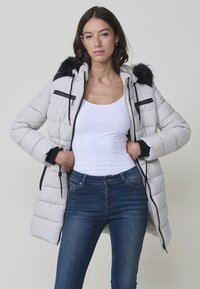 Abrigo acolchado de gris claro con capucha de piel sintética, detalles en negro, bolsillos con cremallera y puños elásticos. Llevado sobre una camiseta blanca sin mangas y vaqueros oscuros.