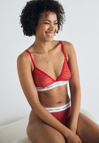 Set di lingerie rosso trasparente composto da un bralette con scollatura a V e un elastico con logo, abbinato a un perizoma, entrambi con motivi a cuore e spalline regolabili.