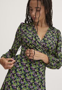 Abito a cascata floreale con sfondo nero e motivi di fiori viola e verdi, scollatura a V e maniche lunghe con polsini elastici.