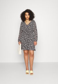 Vestido com estampado floral em cinza escuro, com decote em V, mangas longas, cintura elástica e um ajuste descontraído. Combinado com sandálias amarelas de deslizar e uma mala creme.