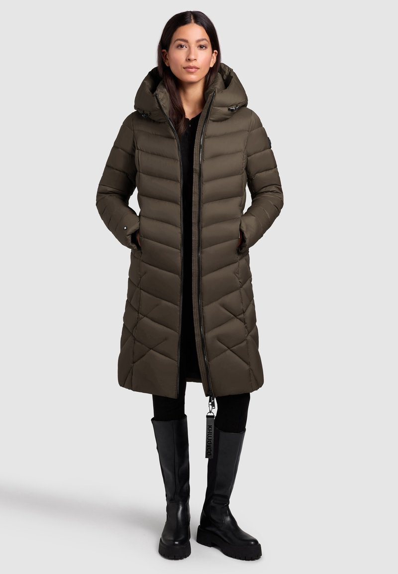 Tumši zaļš pūkains mētelis ar kapuci, ar rāvējslēdzēju priekšpusē, dimanta formas quilted rakstu, sānu kabatām un melnu birku akcentu. Garš, taisns piegriezums.