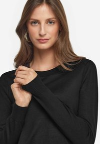 Schwarzer Strickpullover mit Rundhalsausschnitt und gerippten Bündchen. Glatte Textur mit langen Ärmeln, die leicht von den Händen gehalten werden, vor einem neutralen Hintergrund.