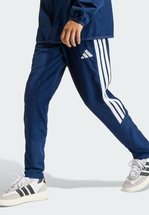 Osoba ubrana w granatowe spodnie dresowe Adidas z białymi paskami oraz białe sneakersy Adidas, idąca naprzód na jednolitym tle.