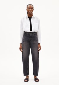 Jeans neri a vita alta con un design a gamba dritta, abbinati a una camicia bianca con bottoni. Il modello si trova di fronte a uno sfondo semplice.