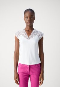 Débardeur blanc avec des manches en dentelle, design en V et texture côtelée. Associé à un pantalon rose, mettant en valeur un contraste de couleurs vibrant.