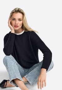 Maglione lavorato a maglia blu navy con texture a coste, abbinato a jeans cropped chiari. La modella è seduta con una mano sul viso, indossando una maglietta bianca sotto.